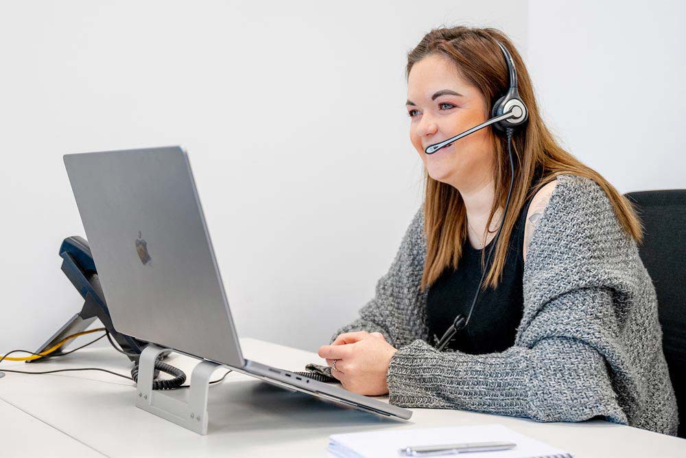Abi from our expert admin team wearing a headset and talking to a customer while taking notes on her MacBook Pro.