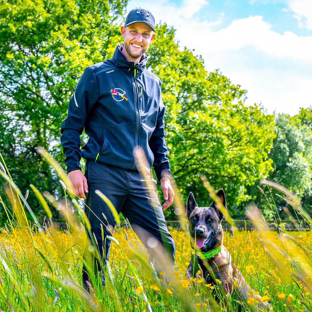 One of our trainers (James) standing in waist-high grass, next to his Belgian malinois named Storm - photo taken at our harrogate training centre at the height of summer